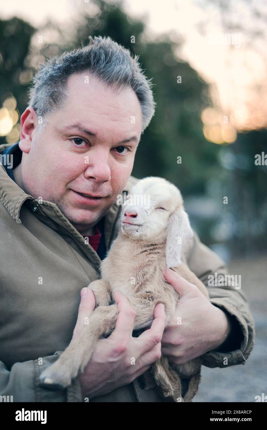 Ein Mann hält ein neugeborenes Ziegenkind in der Nähe, beide schauen in die Kamera Stockfoto