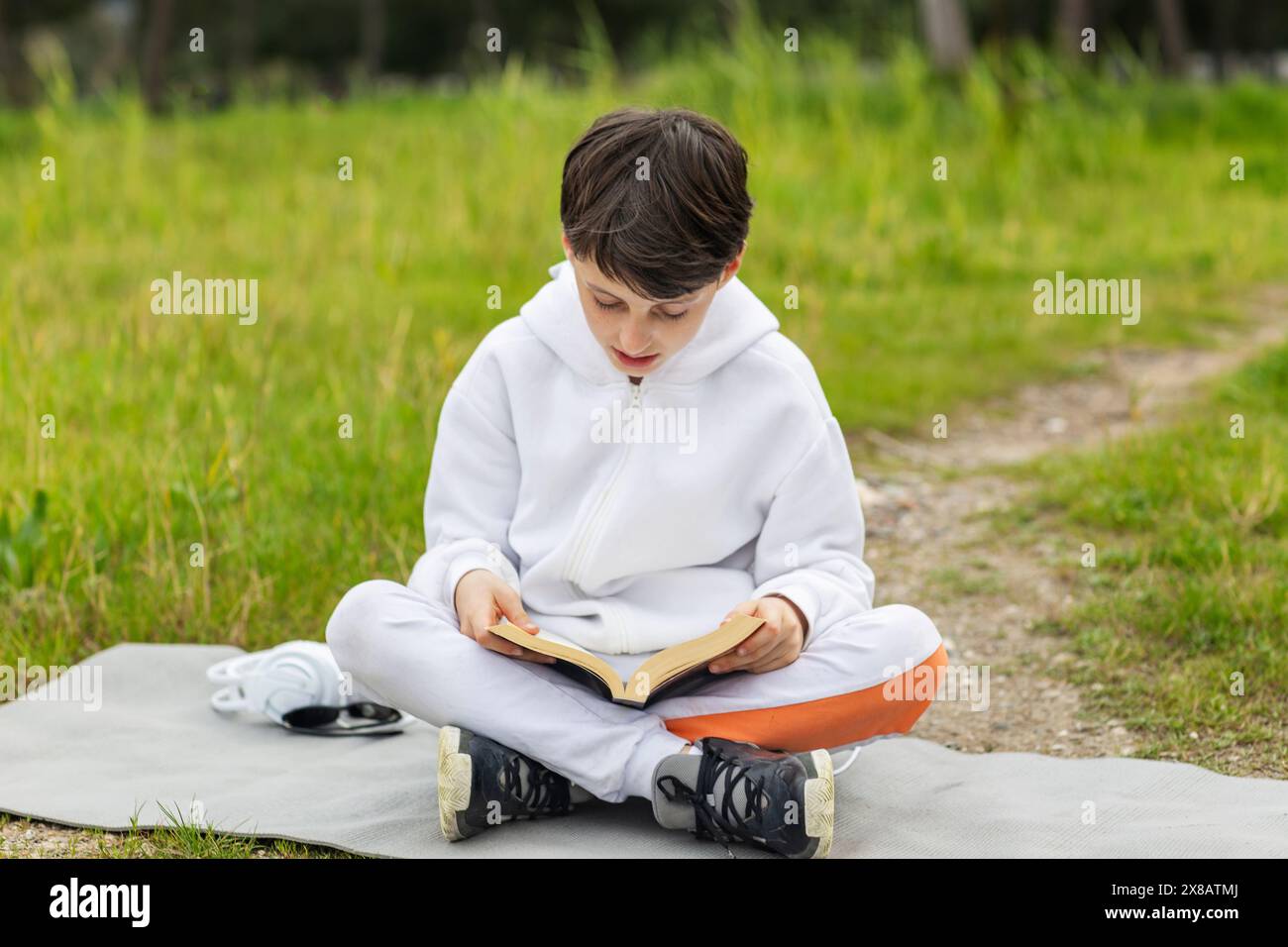 Ein süßer Junge, der ein Buch liest, sitzt auf einer Yogamatte vor dem Waldgrundstück Stockfoto