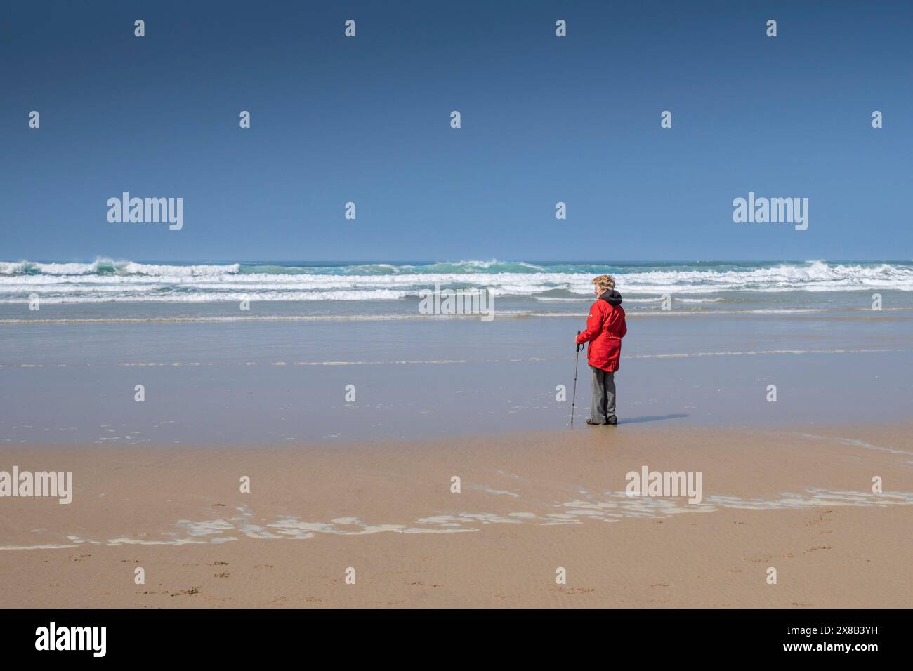 Eine reife Frau, die einen leuchtend roten Mantel trägt und allein am Strand von Mawgan Porth in Cornwall in Großbritannien steht. Stockfoto