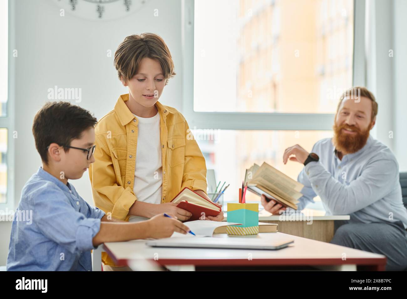 Eine Gruppe von Kindern, die an einem Tisch sitzen und in Bücher vertieft sind, während ein männlicher Lehrer eine lebhafte Diskussion führt. Stockfoto