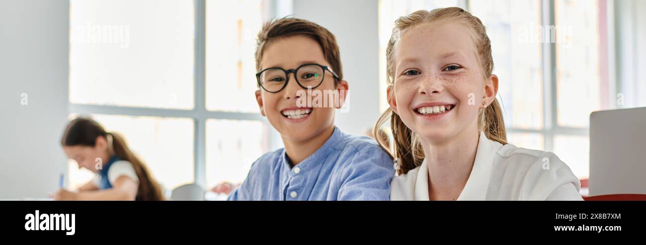 Zwei Kinder, ein Junge und ein Mädchen, sitzen nebeneinander, vertieft in ein Gespräch oder eine Aktivität in einer lebendigen Klassenzimmer-Umgebung. Stockfoto