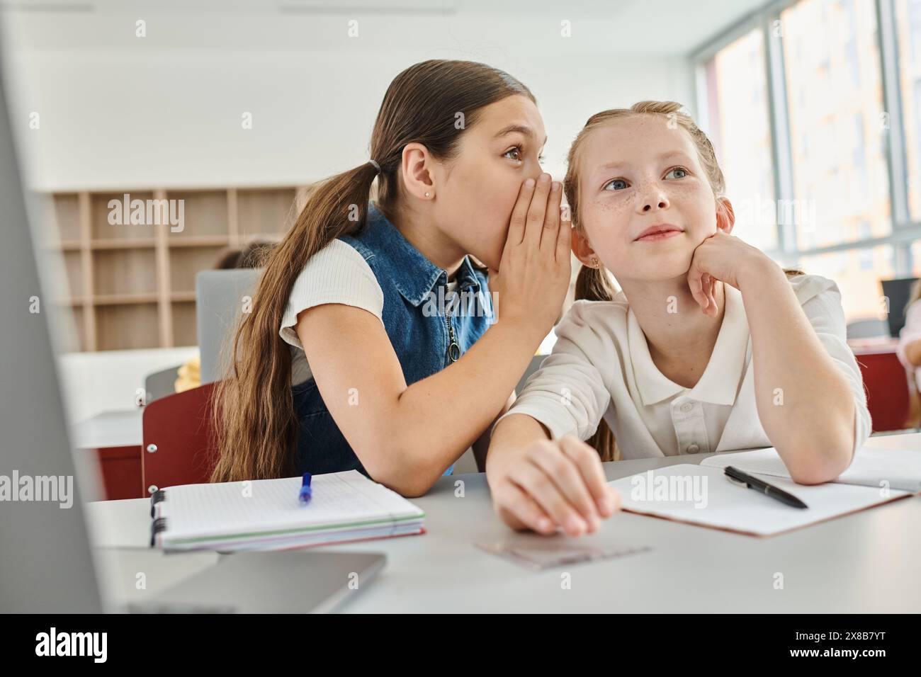 Zwei Mädchen sitzen an einem Tisch und tratschen lebhaft im Klassenzimmer. Stockfoto