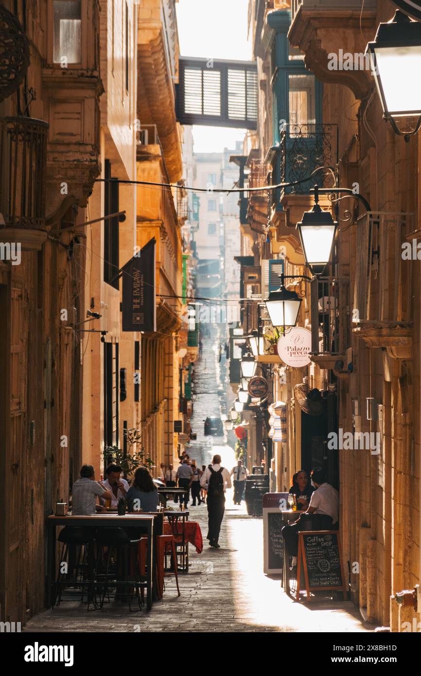 Ein goldener Strahl der Nachmittagssonne durchdringt eine enge Gasse in Valletta, Malta, während die Menschen draußen vor Cafés und Restaurants sitzen Stockfoto