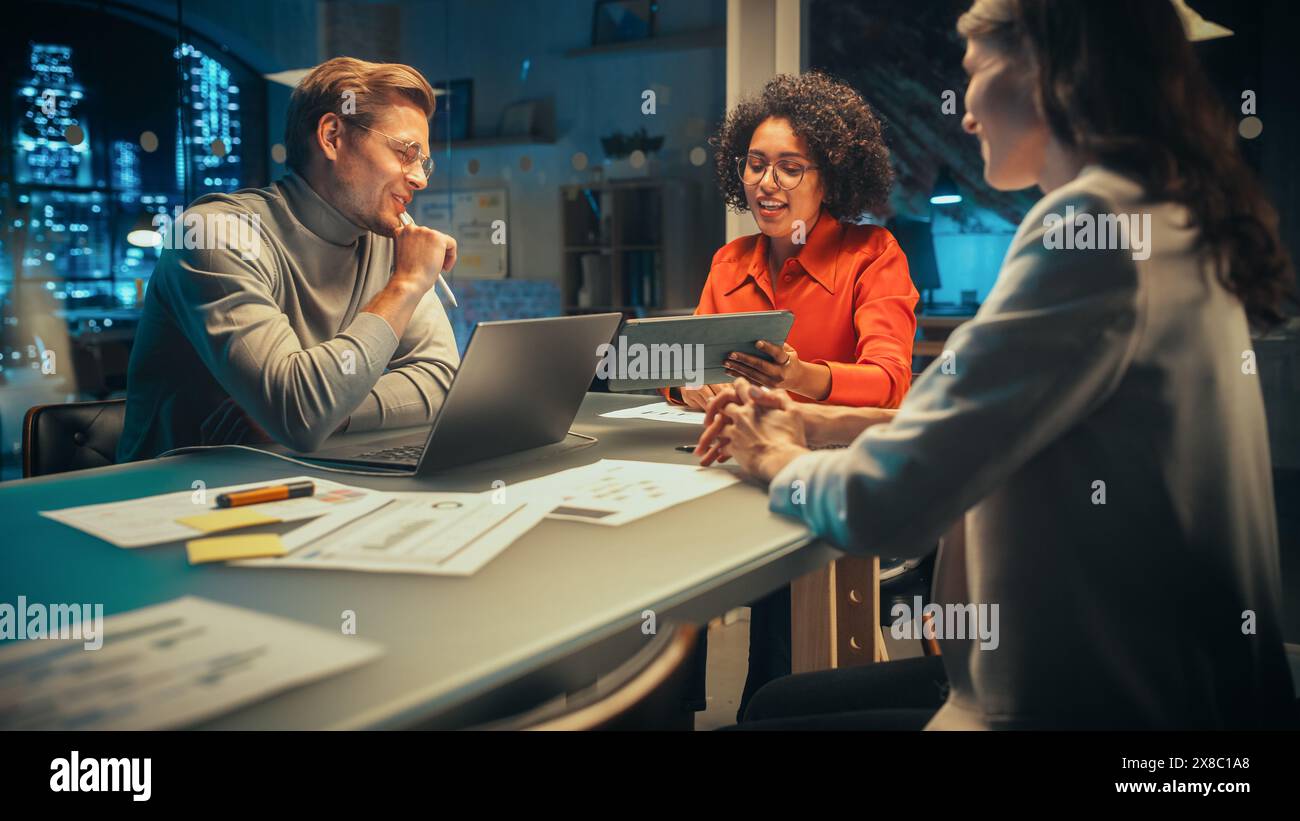 Multikulturell kreative Kollegen, die beiläufig scherzen, eine neue Geschäftschance diskutieren, in einem Besprechungsraum sprechen, ein Notebook und ein Tablet verwenden, um Ideen für das Corporate Marketing zu finden. Stockfoto