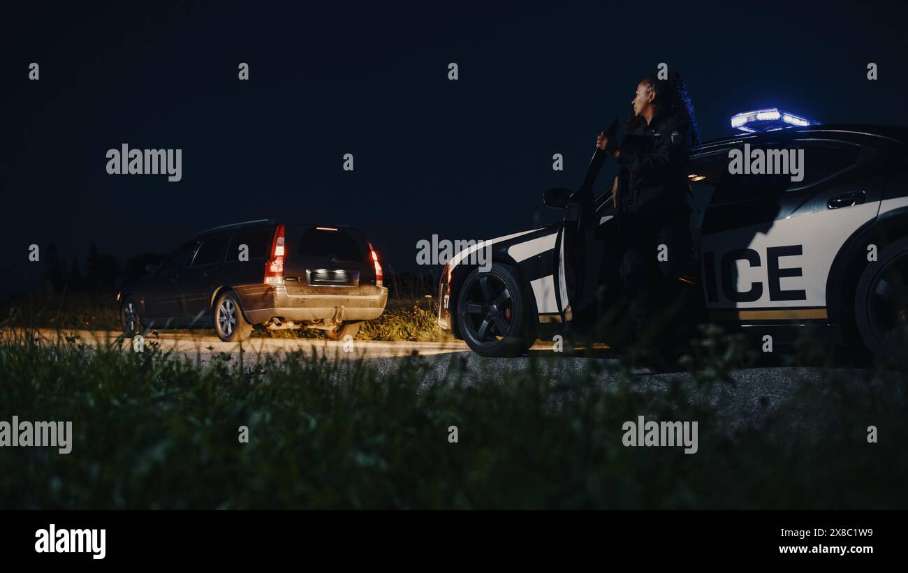 Schwarze Polizistin, die aus dem Patrouillenwagen steigt und auf ein angezogenes Auto zusteuert. Die Polizei antwortete auf einen Anruf von 911 wegen eines verdächtigen Autos, der auf der Straße angehalten wurde, und untersucht die Situation Stockfoto