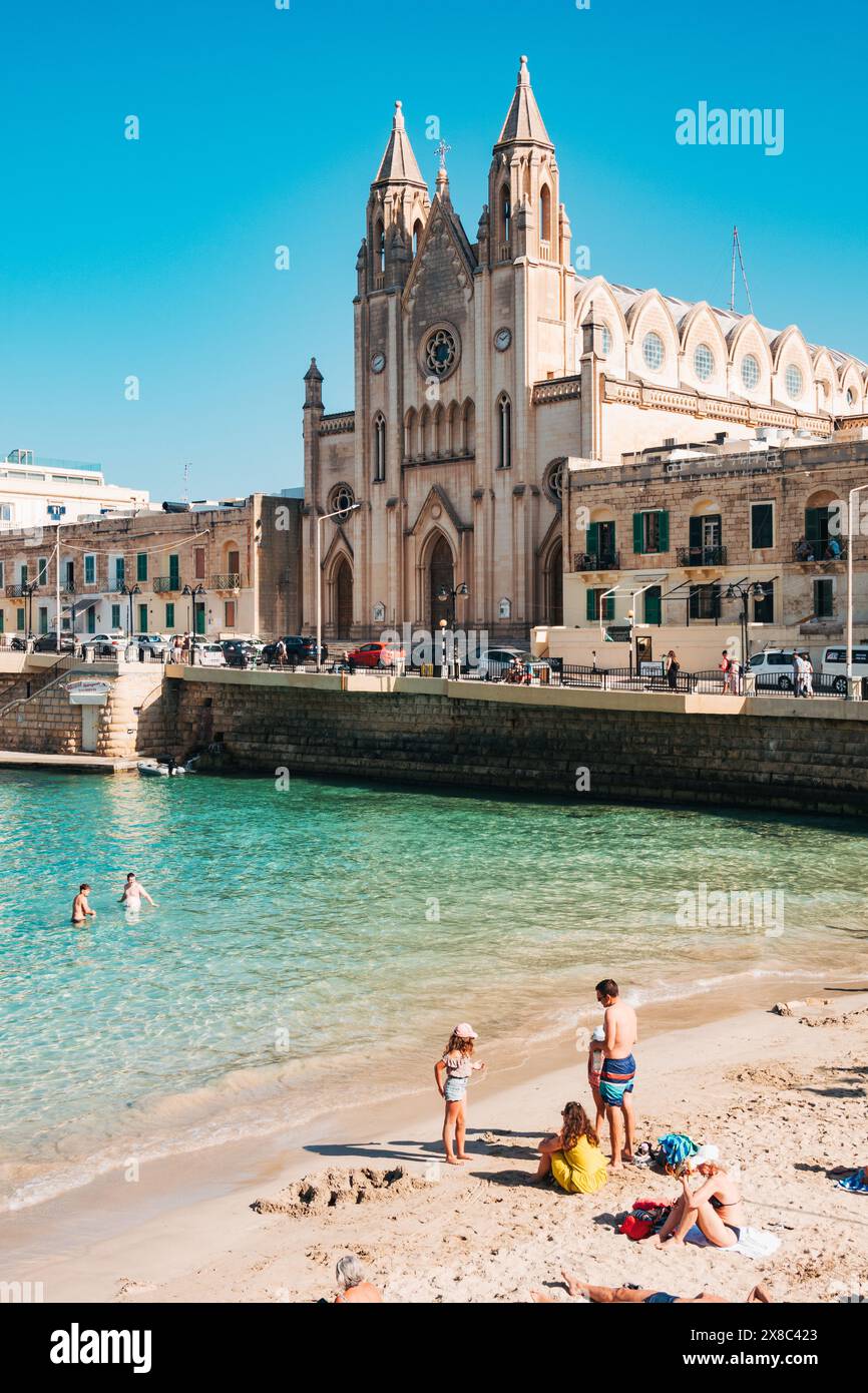 Die Menschen baden im flachen türkisfarbenen Wasser am Balluta Bay Beach in St. Julian's, Malta. Die neogotische Karmeliterkirche ist dahinter zu sehen Stockfoto