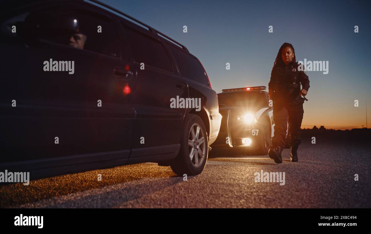 Schwarze Polizistin auf dem Weg zu einem angefahrenen Auto. Fahrer sitzt im Dunkeln. Möglicherweise bewaffneter Fahrer, der auf den Polizisten wartet. Offizier bereitet sich vor, ihre Waffe zu ziehen Stockfoto
