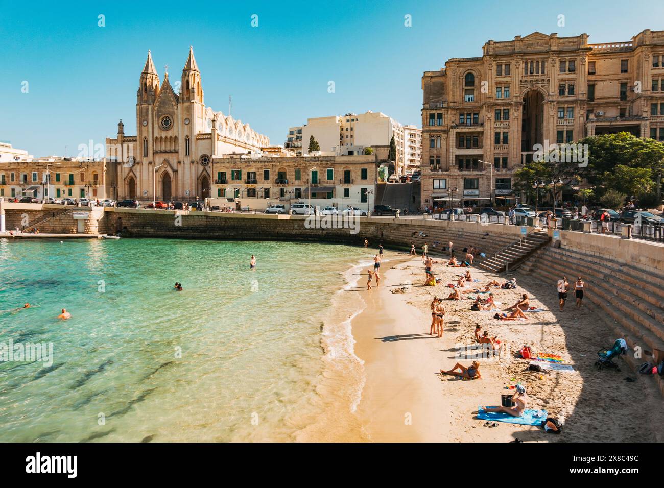 Die Menschen baden im flachen türkisfarbenen Wasser am Balluta Bay Beach in St. Julian's, Malta. Die neogotische Karmeliterkirche ist dahinter zu sehen Stockfoto