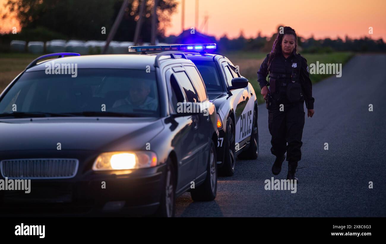 Der Highway Traffic Patrol Car fährt über das Fahrzeug auf der Straße. Der männliche Polizeibeamte kommt zu dem Fahrer und bittet um Lizenz und Registrierung, der schwarze weibliche Kollege deckt ihn ab. Weitwinkelaufnahme In Kinoqualität Bei Dämmerung Stockfoto