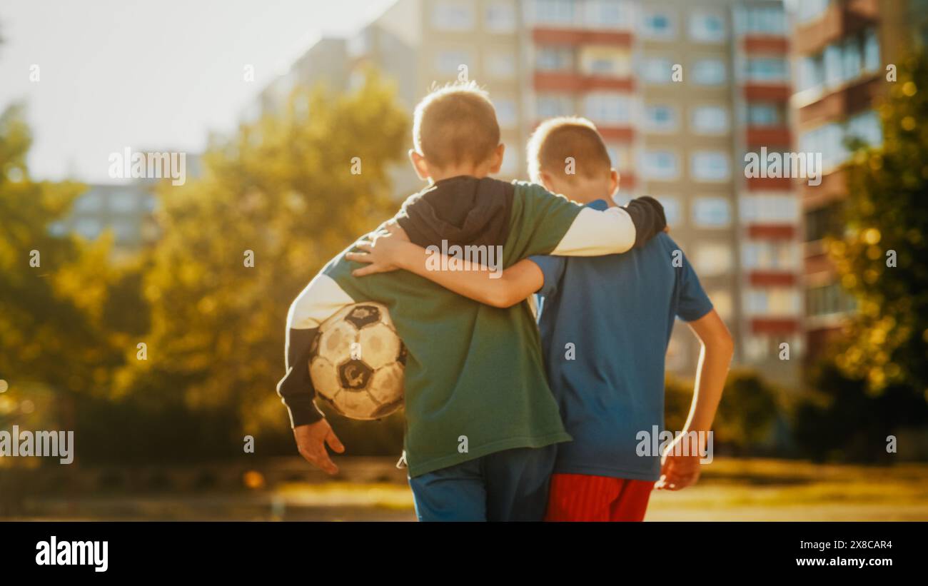 Zwei junge kaukasische Jungen umarmen sich gegenseitig, während sie in Sportkleidung zu einem Fußballplatz in der Nachbarschaft laufen. Fußballspieler, die einen Ball zum Spielen tragen. Das Konzept des Sports, der Kindheit, der Freundschaft. Stockfoto