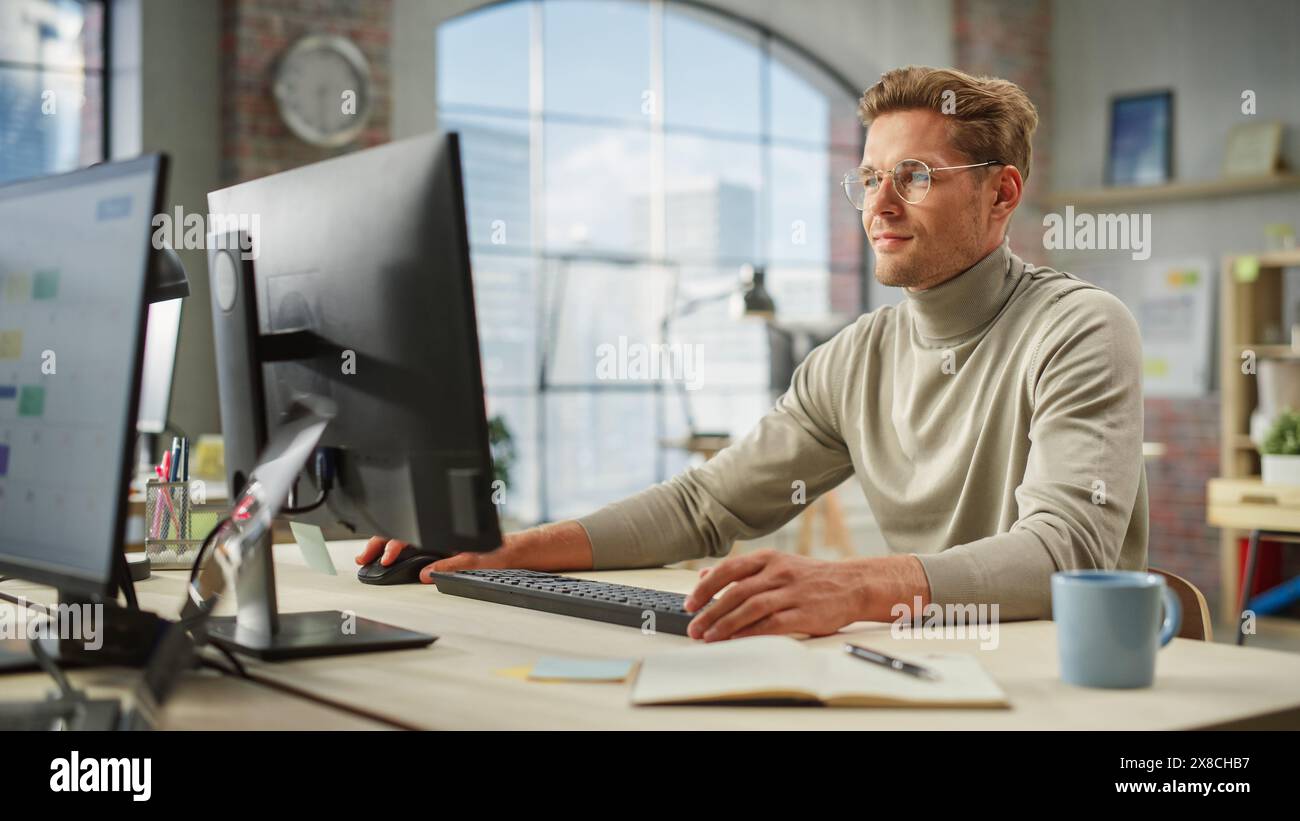 Porträt des kaukasischen kreativen jungen Mannes, der während des Tages im modernen Büro am Computer arbeitet. Der Vorgesetzte Lächelt, Während Er E-Mail-Fragen Von Teammitgliedern Beantwortet. Stockfoto