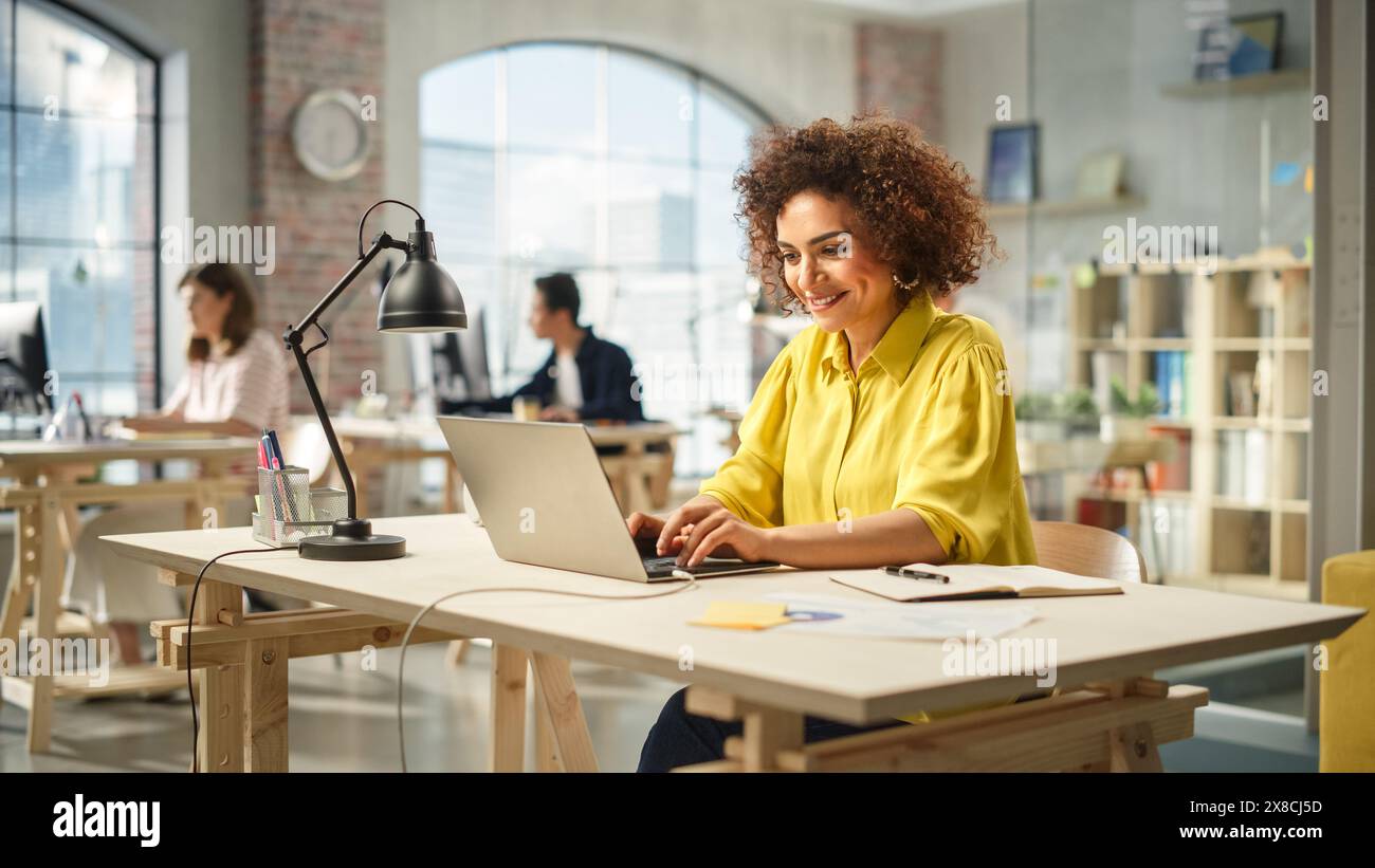 Porträt einer birassischen jungen Frau mit Laptop und Lächeln in einem geräumigen Büro. Technische Supportspezialistin für Frauen, die Probleme mit Software lösen. Kollegen, die im Hintergrund arbeiten. Stockfoto