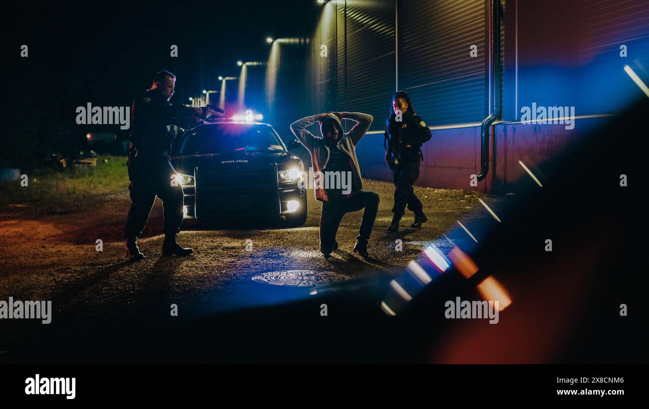 Zwei Polizisten zielen Gub auf den Verdächtigen, befehlen ihm, auf die Knie zu gehen und die Hände zurückzugeben, Verbrecher zu verhaften und anzulegen. Polizisten des Gesetzes legen gefährliche Kriminelle an der Dark City Street an. Kinoaufnahme Stockfoto
