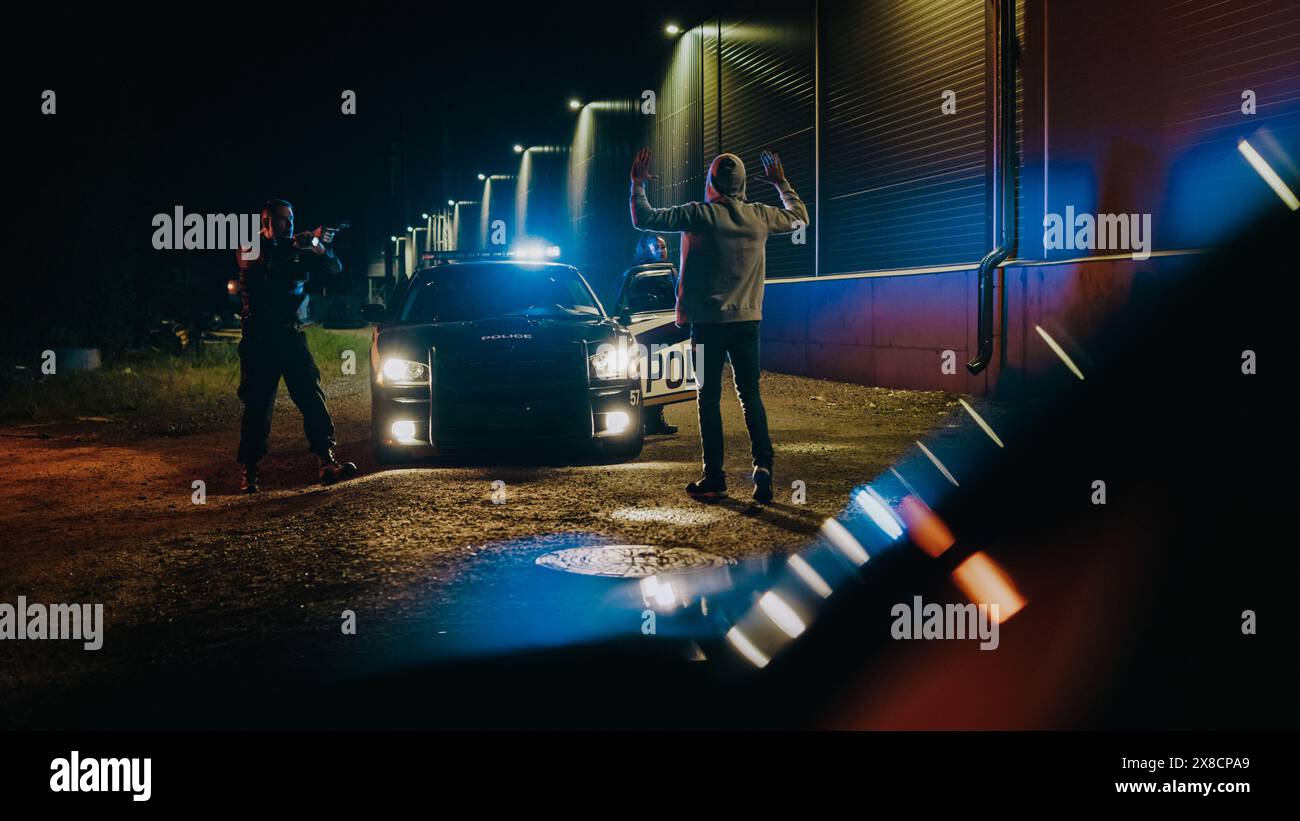 Zwei Polizisten verhaften den Verdächtigen, befehlen ihm, auf die Knie zu gehen und die Hände zurück, Cuff Felon. Polizisten des Gesetzes legen gefährliche Kriminelle an der Dark City Street an. Kinoaufnahme Aus Dem Auto Stockfoto