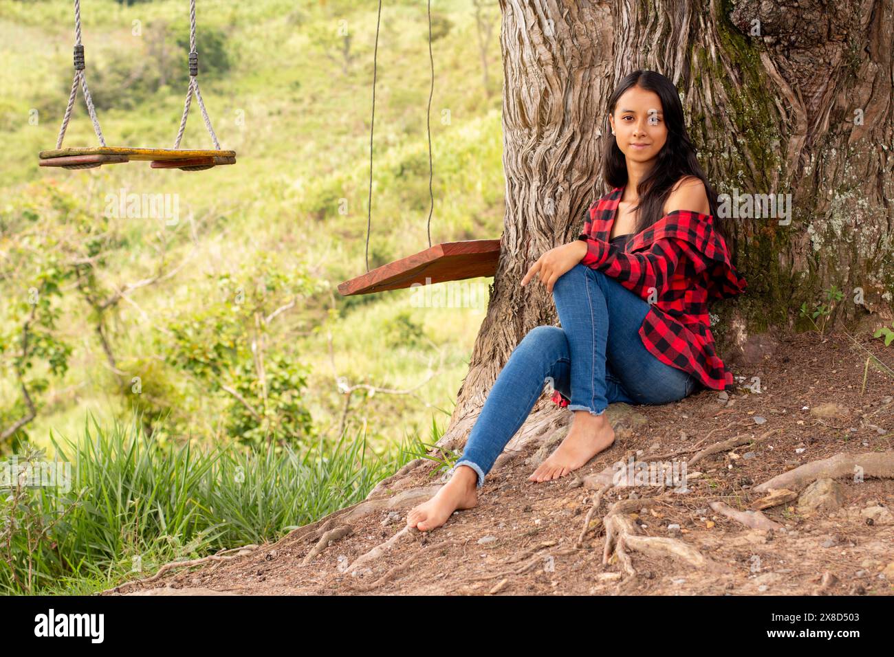 Lebensstil: latina-Frau entspannt sich, während sie barfuß unter einem riesigen Baum in einem Park sitzt Stockfoto