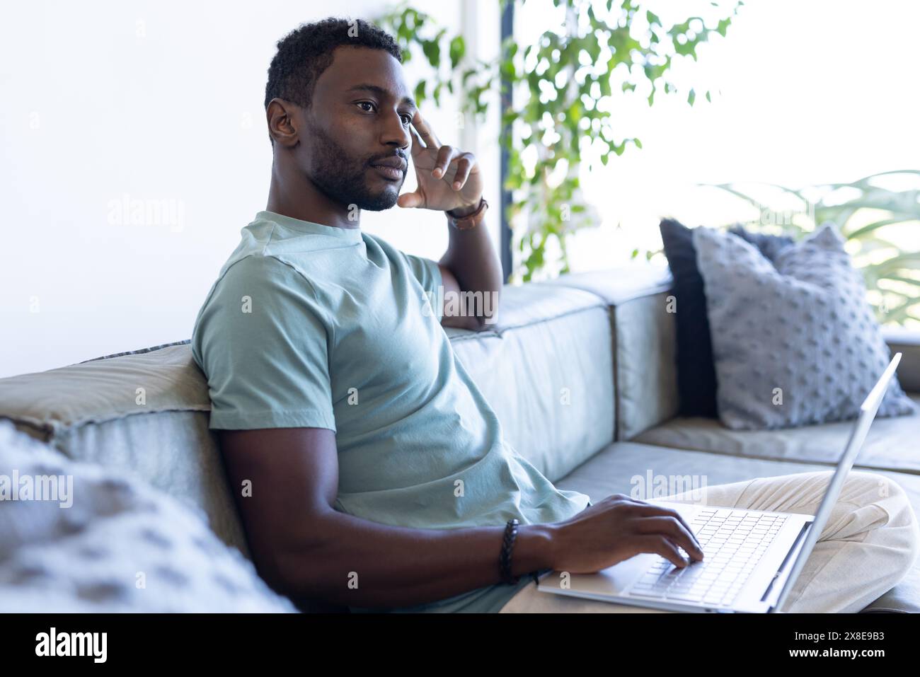 Ein Afroamerikaner zu Hause, der ein hellblaues Hemd trägt und einen Laptop benutzt. Auf der Couch sitzen, einen Anruf tätigen und sich intensiv auf sein Gespräch konzentrieren Stockfoto