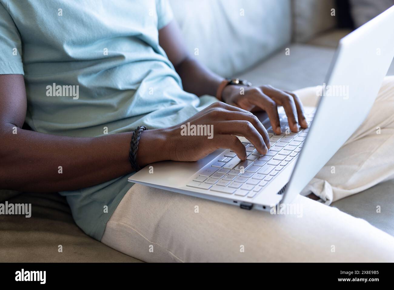 Ein afroamerikanischer Mann, der zu Hause auf dem Laptop tippt und eine Uhr trägt. Mit kurzen schwarzen Haaren und einem hellblauen T-Shirt, das sich intensiv auf das Thema konzentriert Stockfoto