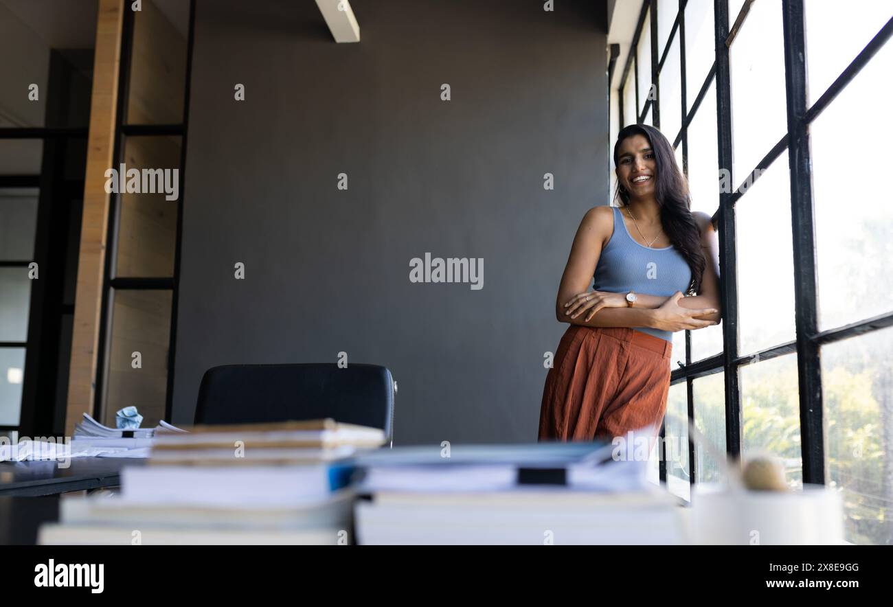 Im modernen Büro steht Indianerin an großen Fenstern, lächelt und sieht entspannt aus, Kopierraum. Sie hat lange schwarze Haare, helle Haut und trägt sie Stockfoto
