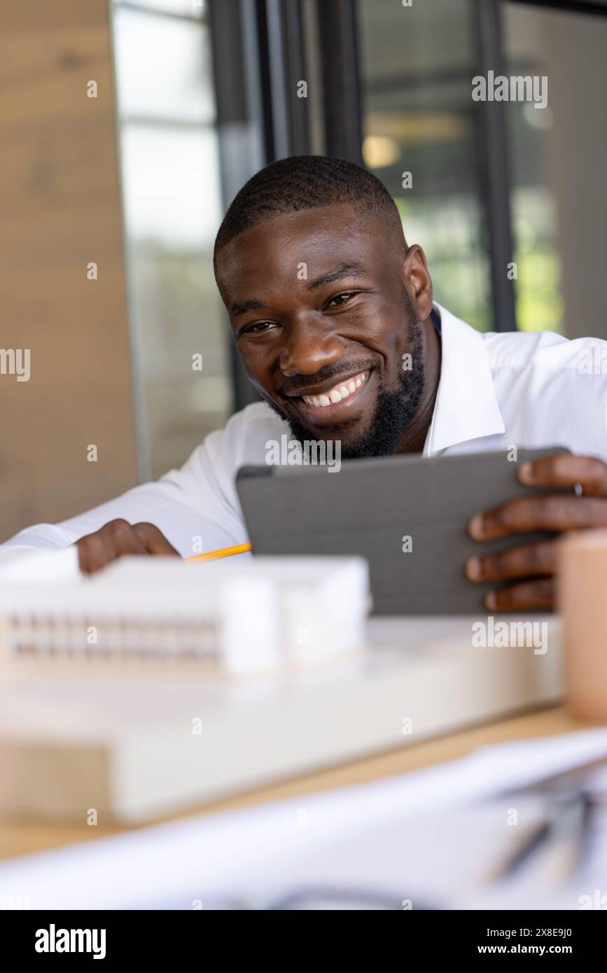 Im modernen Büro lächelt ein junger Afroamerikaner mit kurzen schwarzen Haaren und Bart. Mit weißem Hemd und mit Bürobedarf am Schreibtisch sitzen Stockfoto