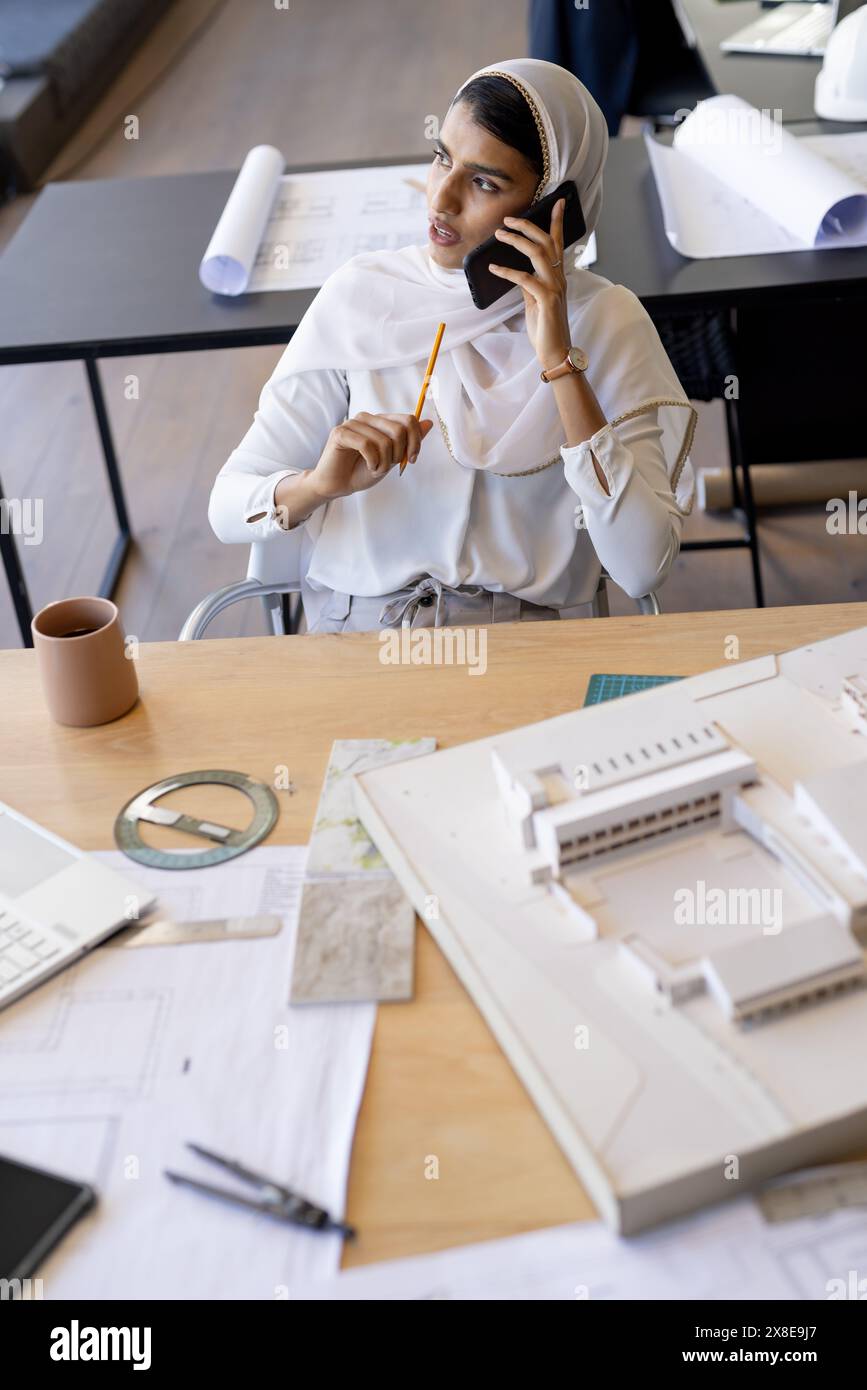 Im modernen Büro, eine junge Indianerin, die Hijab trägt und telefoniert. Sie hält einen Bleistift und sitzt an einem Schreibtisch mit Architekturzeichnungen und einem M Stockfoto