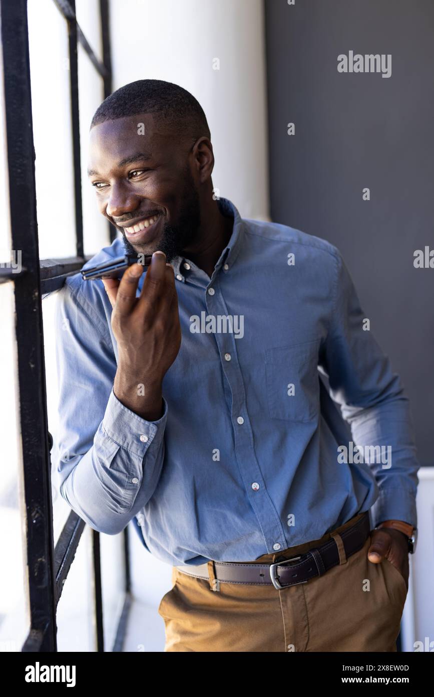 Im modernen Büro stand ein junger Afroamerikaner am Fenster, lächelte und redete. Kurzes schwarzes Haar, beschnittener Bart, blaues Hemd mit Button-up und braun Stockfoto
