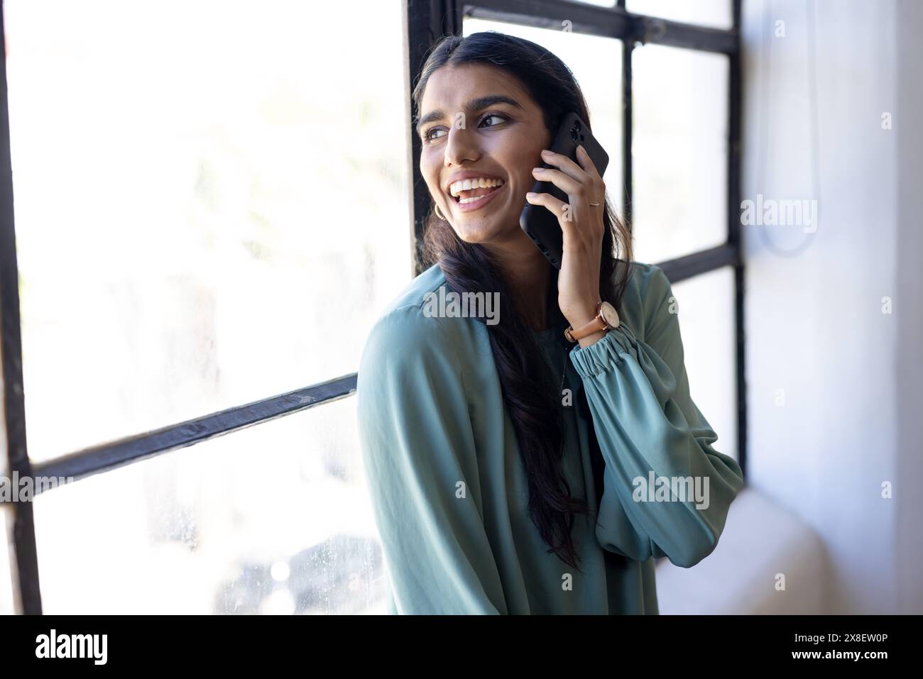indische Frau mit langen schwarzen Haaren und heller Haut spricht im modernen Büro auf Smartphone. Tragen Sie hellgrüne Bluse und sehen Sie zu, lächeln Sie, während Sie nach draußen schauen Stockfoto