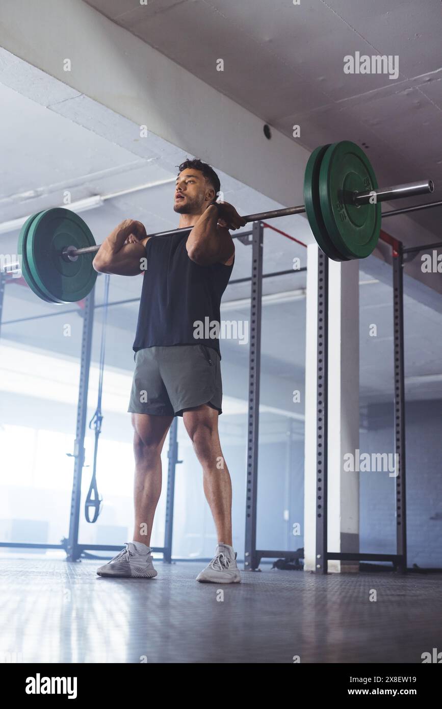 Im Fitnessstudio hebt ein starker und fitter birassischer Mann die Langhantel und konzentriert sich auf sein Workout. Umgeben von Gewichthängen und Fitnessgeräten, schaffen Sie ein spezielles T Stockfoto