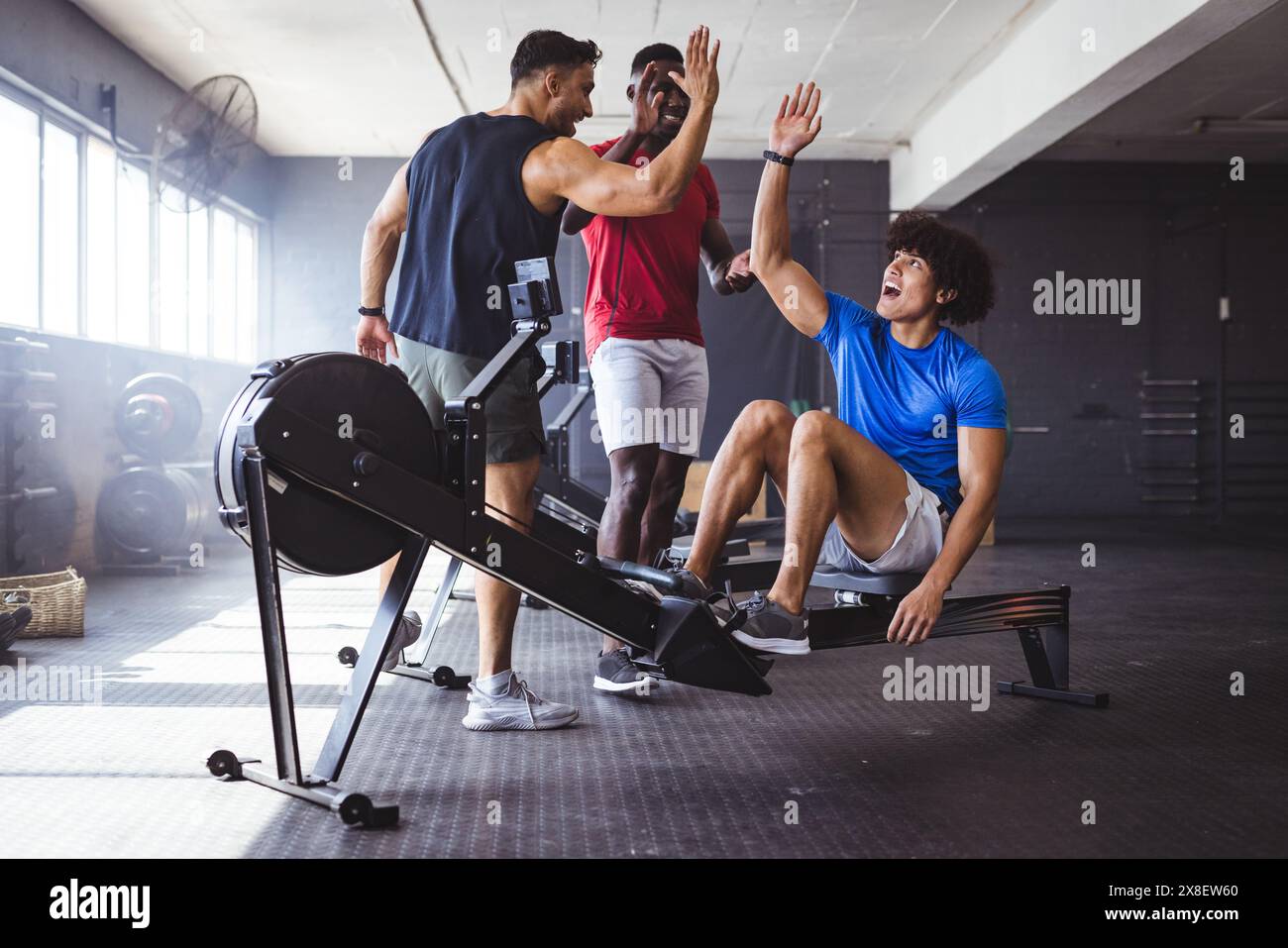 Im Fitnessstudio sind drei junge, fitte, vielfältige Männer in der Nähe des Rudergeräts High-Five. Sie befinden sich in einem geräumigen, gut beleuchteten Fitnessraum mit großen Fenstern und verschiedenen Workouts Stockfoto