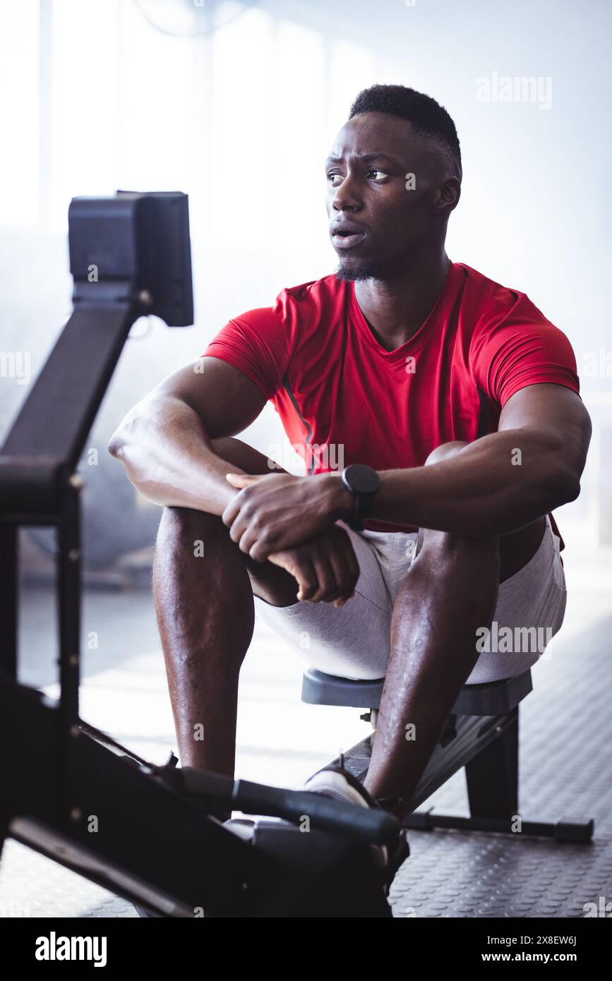 Afroamerikaner, Mitte Zwanziger, auf Rudermaschine, Blick auf den Bildschirm im Fitnessstudio. Fitnessgeräte und helles Tageslicht sorgen für ein energiegeladenes Workout Stockfoto