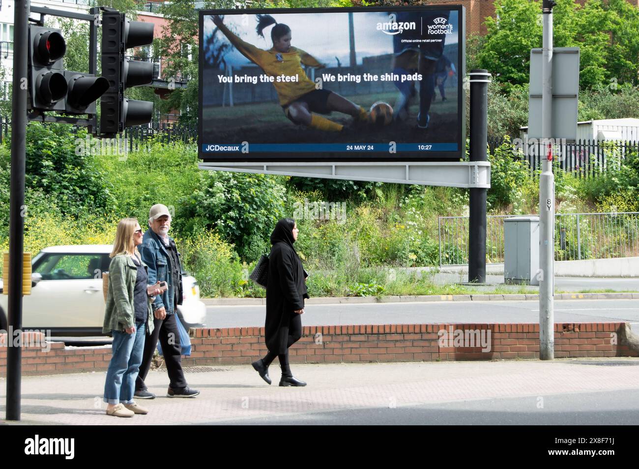 Drei Personen auf der Insel einer Fußgängerüberquerung in kingston, surrey, unterhalb einer digitalen Werbetafel, die amazon im fernsehen über Frauenfußball anpreist Stockfoto