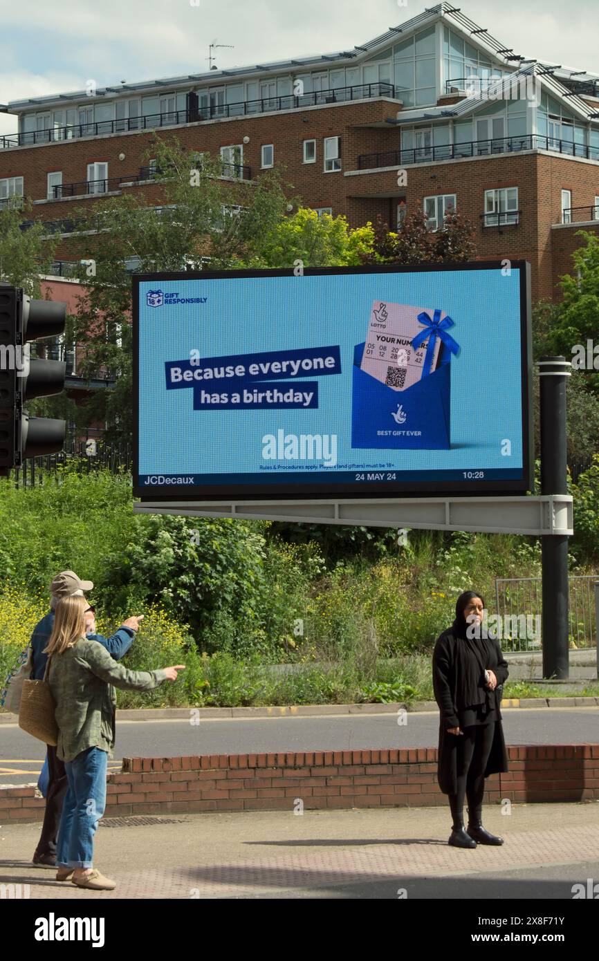 Drei Personen auf der Insel eines Fußgängerübergangs in kingston, surrey, england, unter einer digitalen Werbetafel, die für die britische nationale Lotterie wirbt Stockfoto