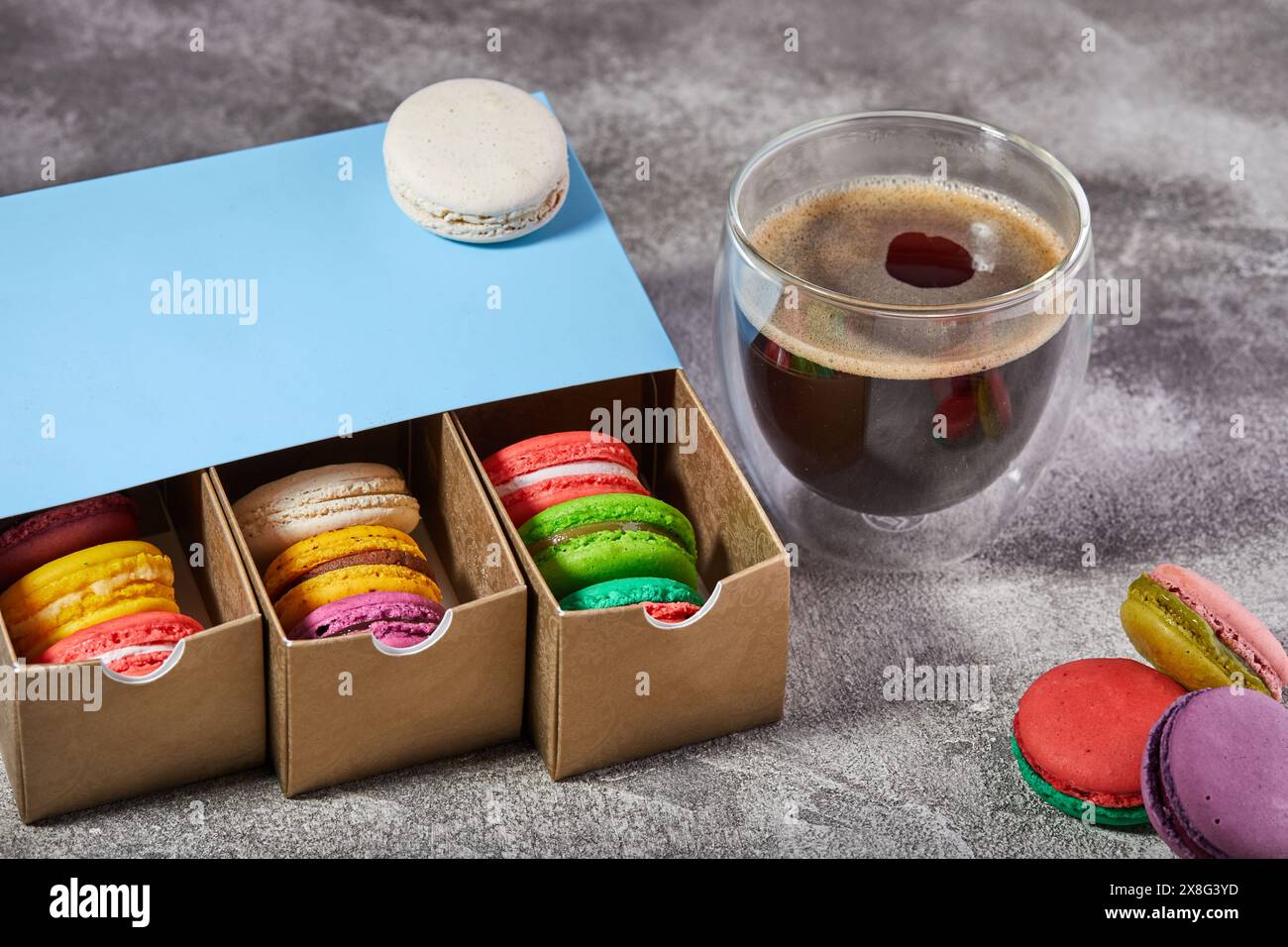 Ein Tisch mit einer Schachtel mit bunten Macarons neben einer dampfenden Tasse Kaffee. Die perfekte Kombination aus süßen Leckereien und einem warmen Getränk Stockfoto