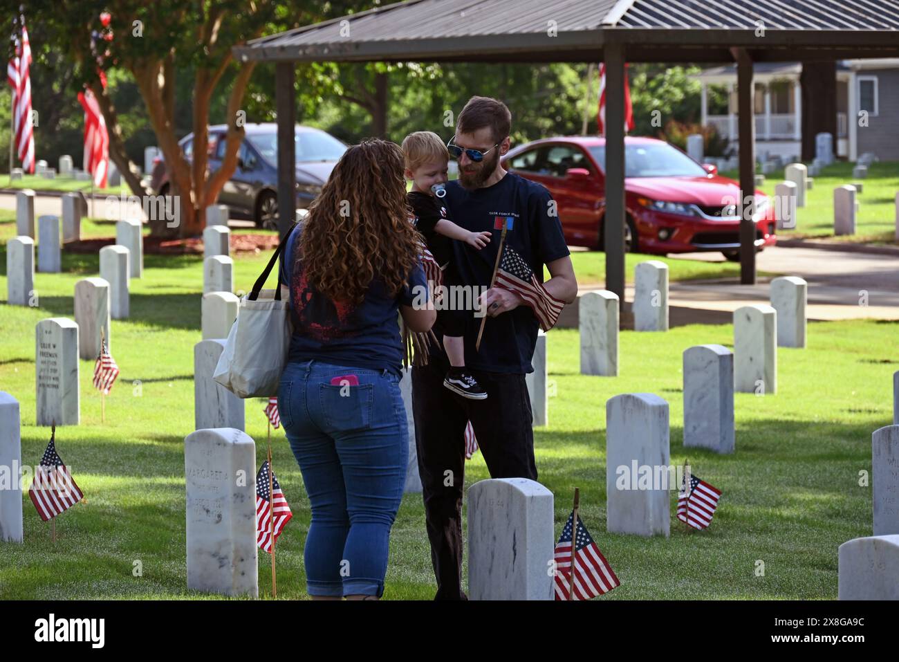 Raleigh, NC, USA, 25. Mai 2024; eine junge Familie schließt sich freiwilligen Helfern an, die Fahnen an den 6.000 Grabsteinen auf dem Raleigh National Cemetery vor dem Memorial Day-Feiertag platzieren. Die jährliche Veranstaltung, die von der Raleigh American Legion organisiert wird, ehrt die amerikanischen Männer und Frauen, die in Friedenszeiten und Krieg im US-Militär gedient haben. Der Raleigh National Cemetery umfasst die Grabstätten von Soldaten aus dem Bürgerkrieg bis zu den Kriegen im Irak und Afghanistan. Credit D Guest Smith / Alamy Live News Stockfoto