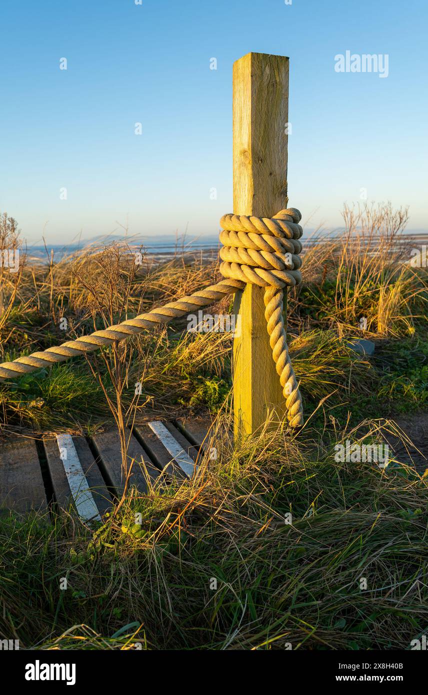 Ein rustikaler Holzpfosten mit dickem Seil in einem grasbewachsenen Feld unter klarem blauem Himmel während der goldenen Stunde. Stockfoto