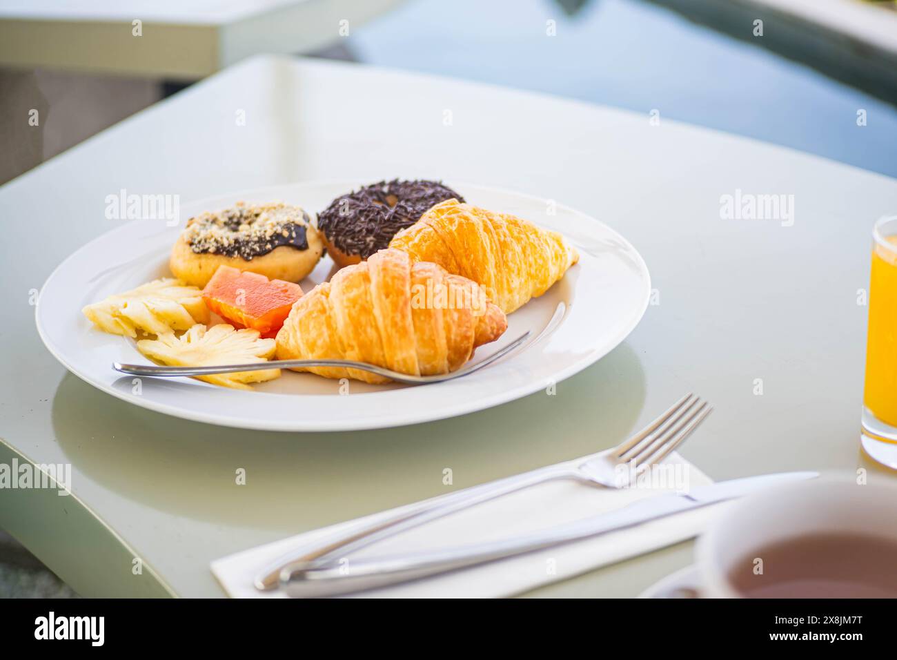 Ein Frühstücksteller mit Croissants, Donuts, Ananasscheiben und einem Stück Lachs, begleitet von einem Glas Orangensaft und einer Tasse Kaffee auf einer Ta Stockfoto