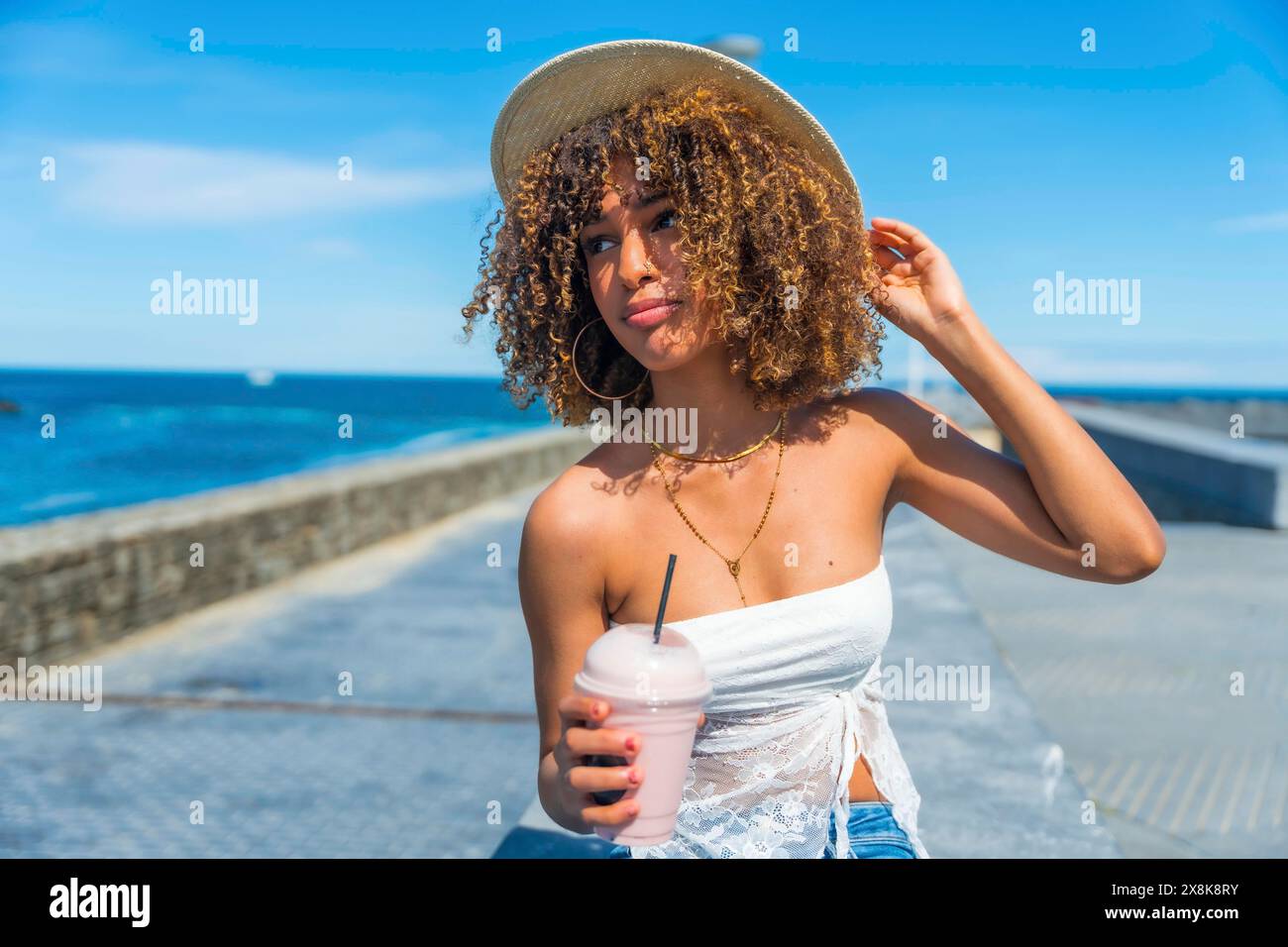 Modische junge lateinfrau mit lockigen Haaren, die einen Hut trägt, der Saft zum Mitnehmen am Meer trinkt Stockfoto