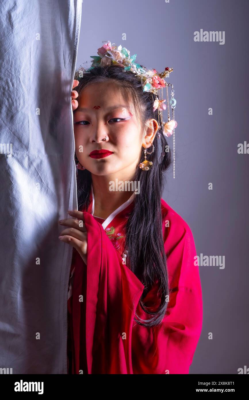 Ein junges Mädchen in einem roten Kleid mit einem Blumenhaarband steht hinter einem Vorhang. Sie hat roten Lippenstift und ihre Augen sind geschlossen. Begriff des Mysteriums und Stockfoto