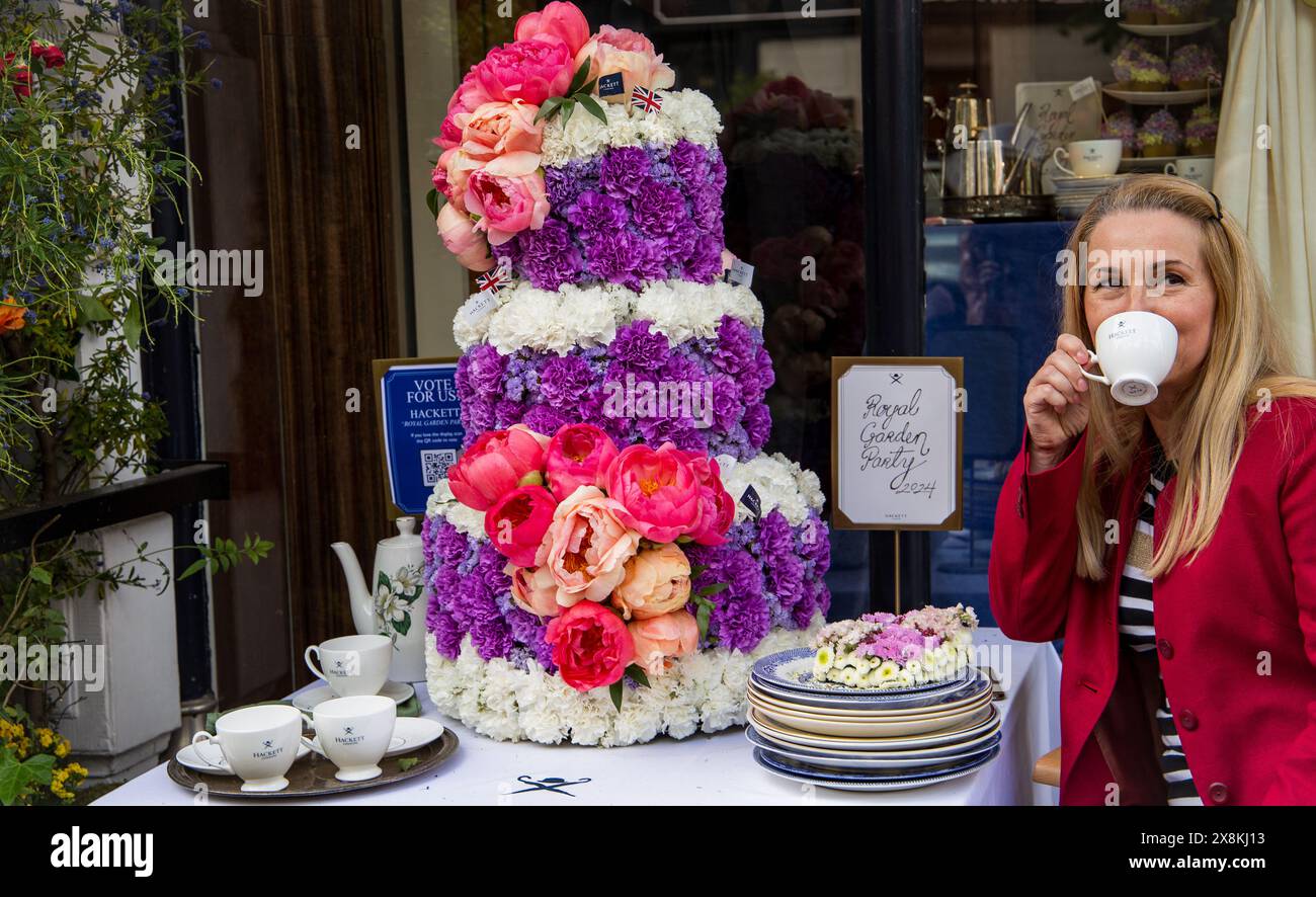 Frau in roter Jacke trinkt Tee neben einer gestuften Blumenkuchen-Ausstellung in Chelsea in Blüte außerhalb des Ladens. Royal Garden Party Thema. Stockfoto