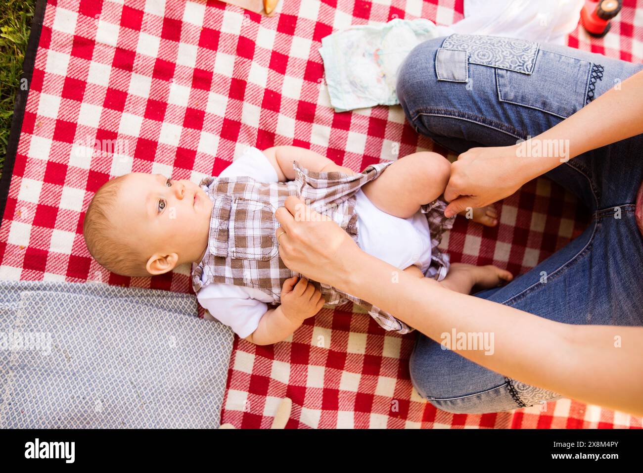 Das Baby liegt auf einer Picknickdecke während des Picknickens in der Gruppe im Park. Mutter wechselt die Windel. Stockfoto