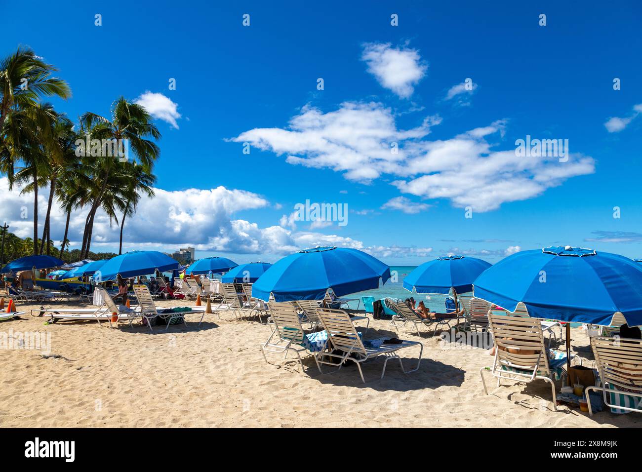 Touristen entspannen sich auf Chaiselongen mit blauen Sonnenschirmen am sonnigen Waikiki Beach in Honolulu, Hawaii. Stockfoto