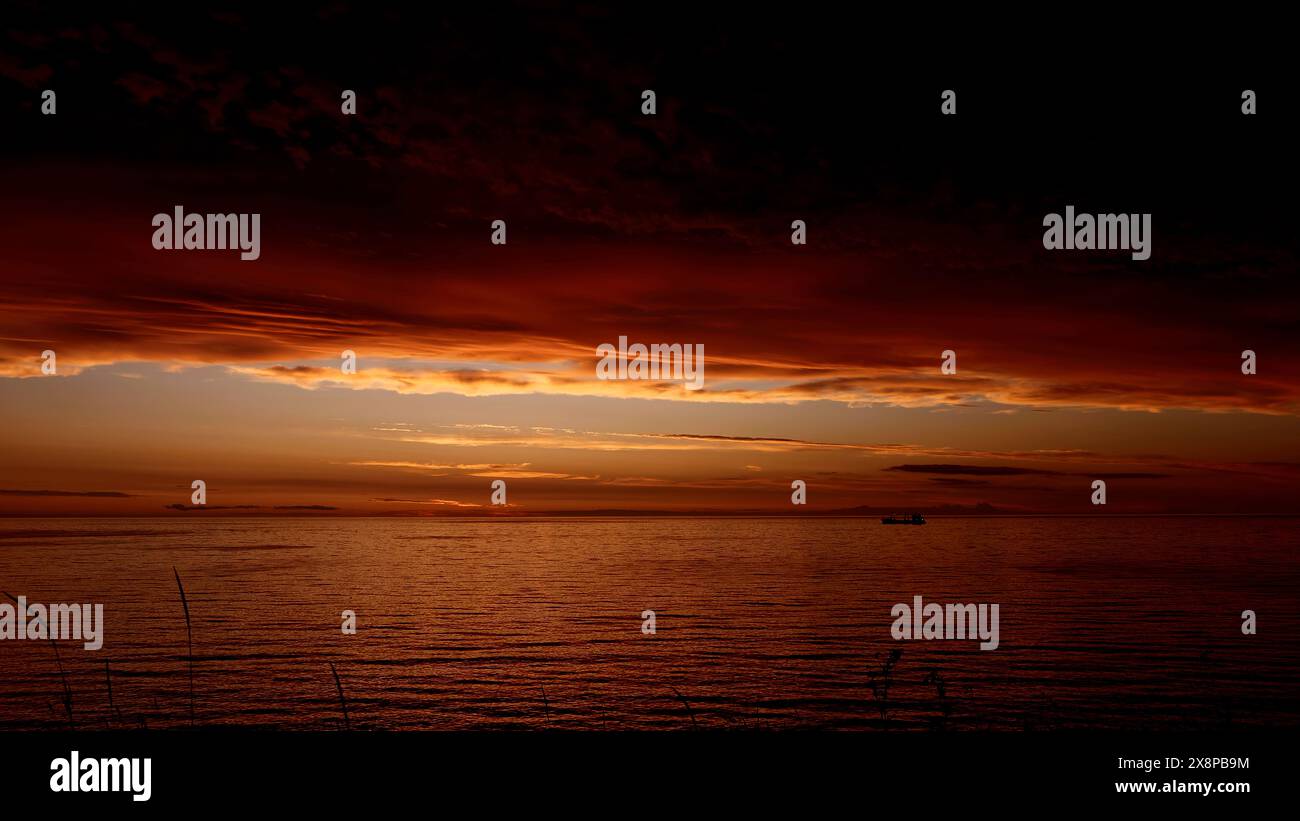 Märchenhafter atemberaubender Sonnenuntergang über dem plätschernden Fluss mit einem schwimmenden Boot. Clip. Dunkelroter und oranger Himmel, schwere Wolken über Wasser. Stockfoto