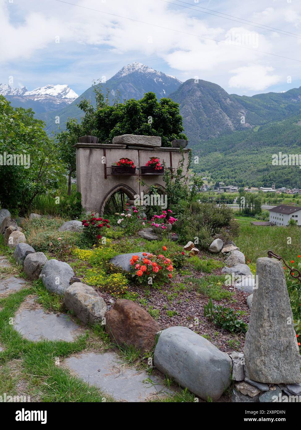 Schrein am Straßenrand mit Blumen im Frühling mit alpinen Bergen dahinter in NUS im Aostatal NW Italien. Mai 2024 Stockfoto