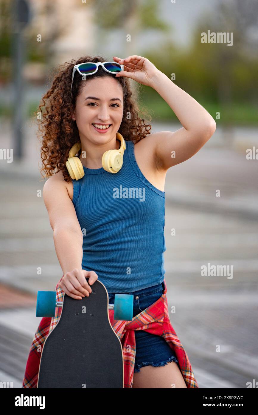Eine fröhliche junge Frau mit lockigem Haar, Sonnenbrille und Kopfhörer, hält ein Skateboard in einem urbanen Park. Stockfoto