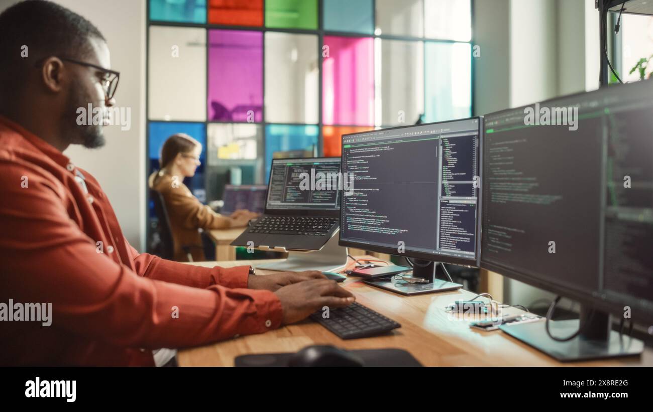 Afroamerikanischer Mann, der Code auf einem Desktop-Computer mit mehreren Monitoren und einem Laptop im Kreativbüro schreibt. Professioneller Datenwissenschaftler mit Software zur Analyse von Informationen aus dem Internet. Stockfoto
