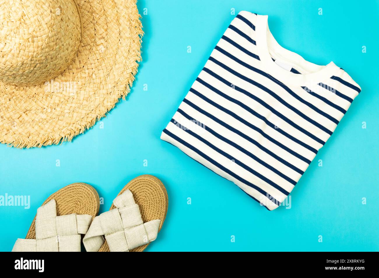 Blick von oben auf Strohhut, Hausschuhe und gestreiftes T-Shirt auf blauem Hintergrund. Sommermode, Urlaubs- und Strandtapete. Kopierraum, flach. Stockfoto