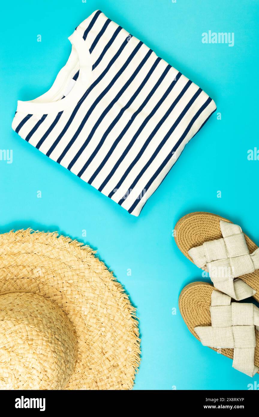 Blick von oben auf Strohhut, Hausschuhe und gestreiftes T-Shirt auf blauem Hintergrund. Sommermode, Urlaubs- und Strandtapete. Kopierraum, flach. Stockfoto