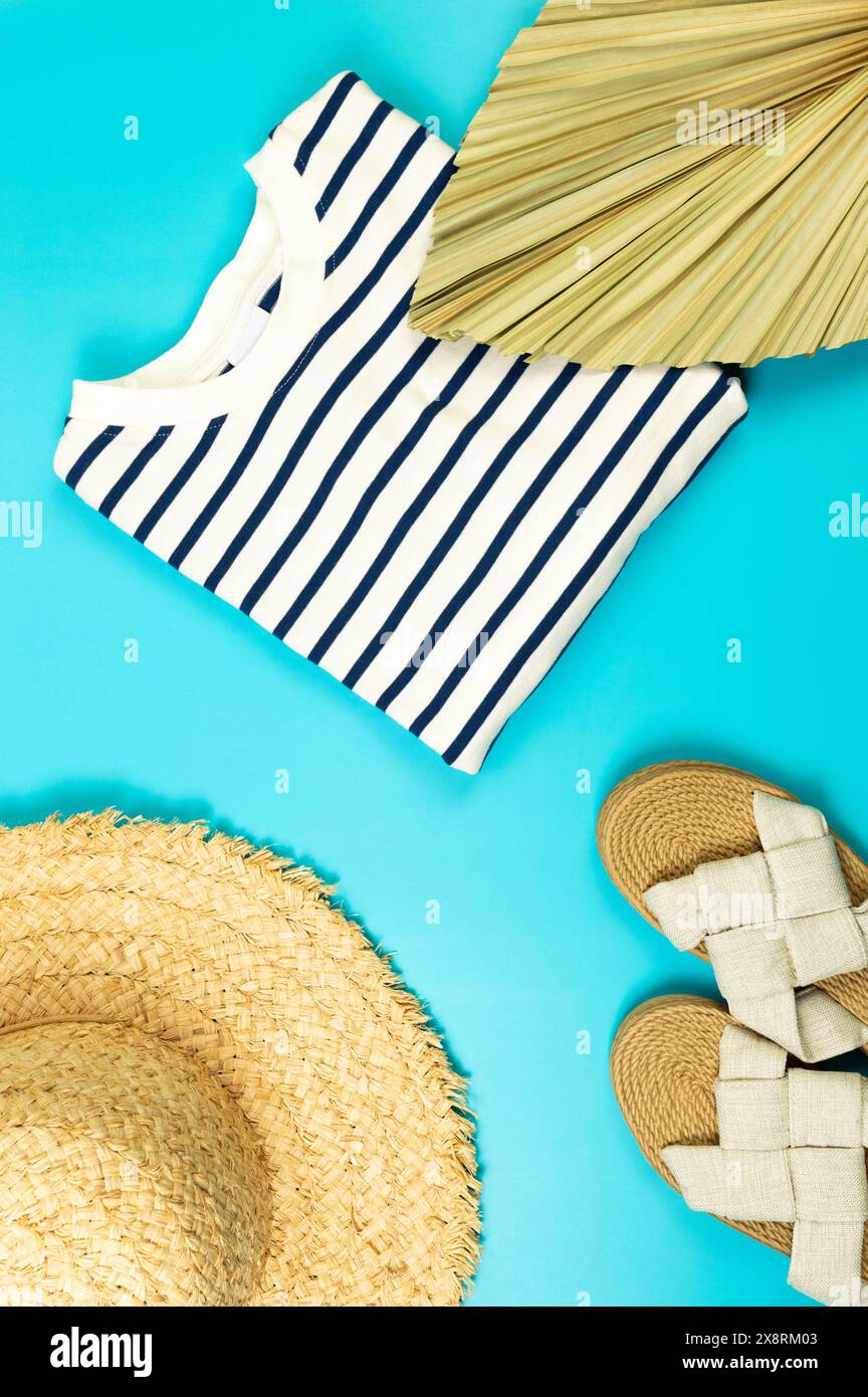 Blick von oben auf Strohhut, Hausschuhe, getrocknete Palmblätter und gestreiftes T-Shirt auf blauem Hintergrund. Sommermode, Urlaubs- und Strandtapete. Kopierbereich, fla Stockfoto