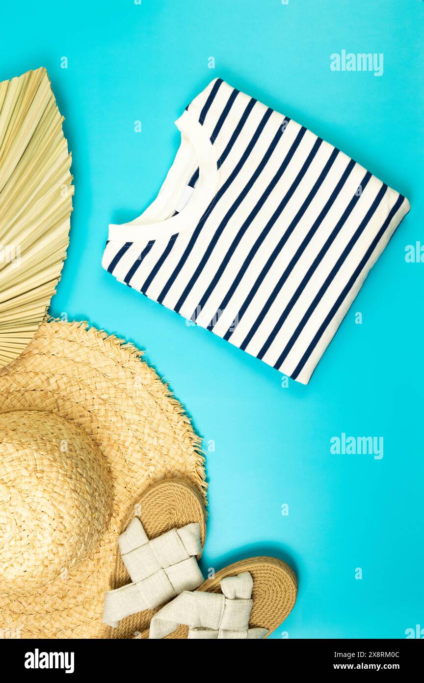 Blick von oben auf Strohhut, Hausschuhe, getrocknete Palmblätter und gestreiftes T-Shirt auf blauem Hintergrund. Sommermode, Urlaubs- und Strandtapete. Kopierbereich, fla Stockfoto