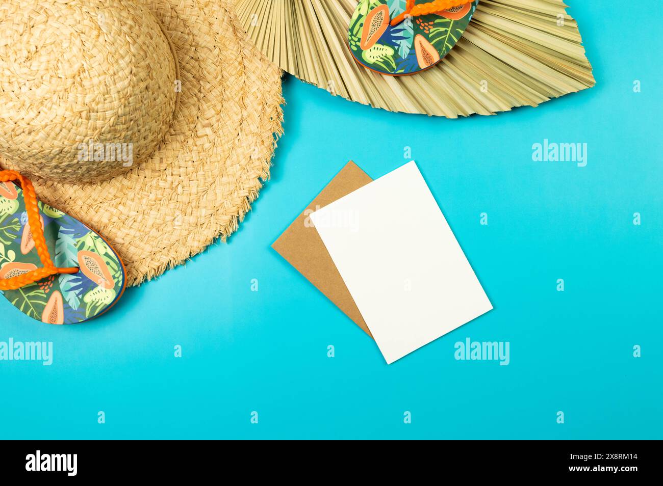 Draufsicht auf Strohhut, getrocknete Palmblätter, weiße Karte und Hausschuhe auf blauem Hintergrund. Sommermode, Urlaubs- und Strandtapete. Kopierraum, flach. Stockfoto