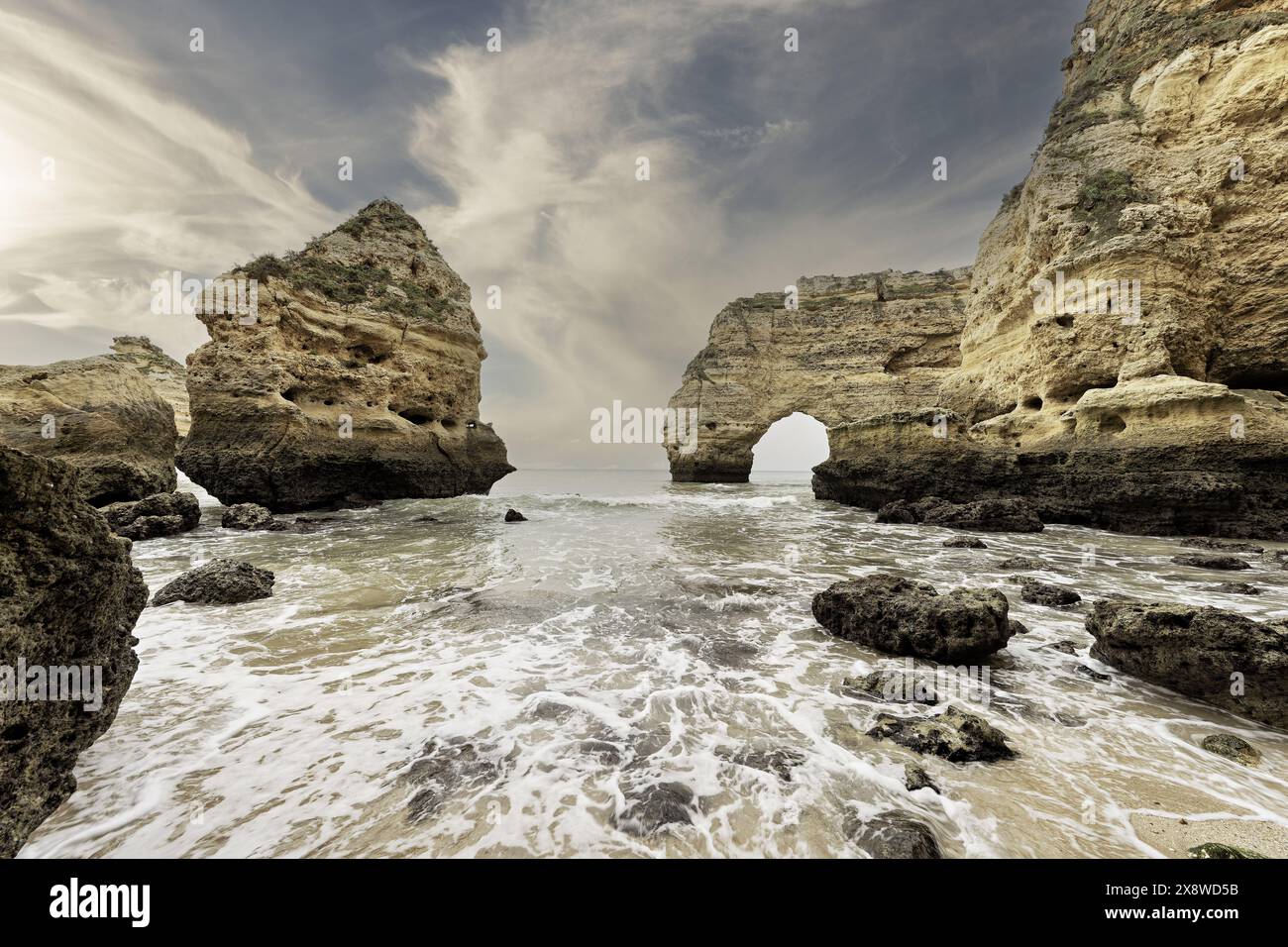 Ein Felsvorsprung am Marinha Beach, Portugal, mit einer Silhouette vor dem sanften Licht eines bewölkten Himmels. Wellen schlängeln sanft am Ufer und schaffen eine ruhige Küste Stockfoto